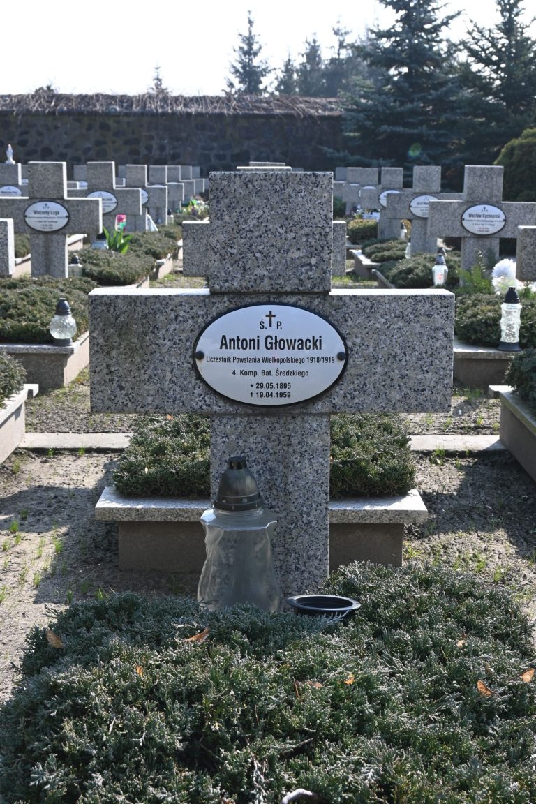 Kamienny grób z krzyżem i owalną tabliczką „Antoni Głowacki”; przed krzyżem szary szklany znicz na czarnym spodku, wokół niski jałowiec, w tle inne nagrobki.