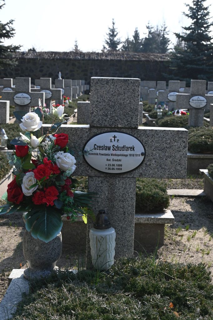 Kamienny krzyż nagrobny z owalną tabliczką „Czesław Szkudlarek”; obok krzyża duży bukiet czerwonych i białych róż w zielonym wazonie oraz biały lampion, w tle rędy krzyży i kamienny mur.