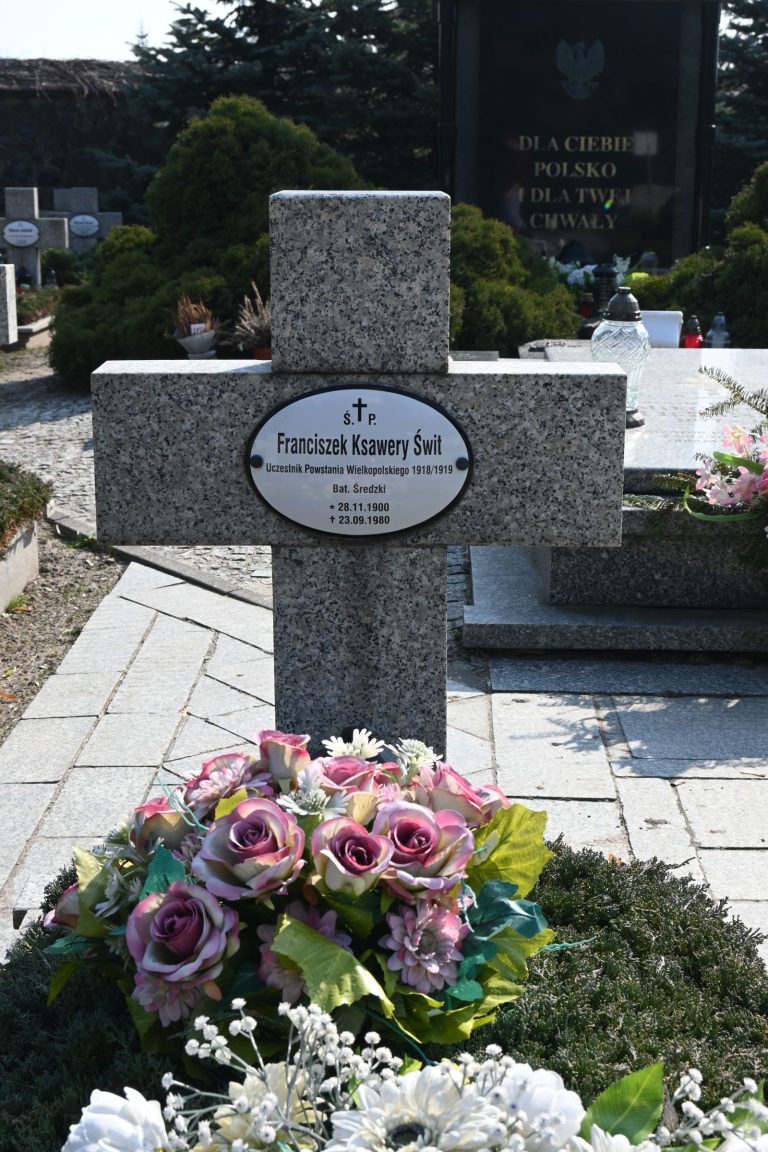 Kamienny krzyż nagrobny z owalną tabliczką "Franciszek Ksawery Świt"; u stóp duża kompozycja różowych róż i białych drobnych kwiatów, w tle brukowana alejka i czarny pomnik z orłem.