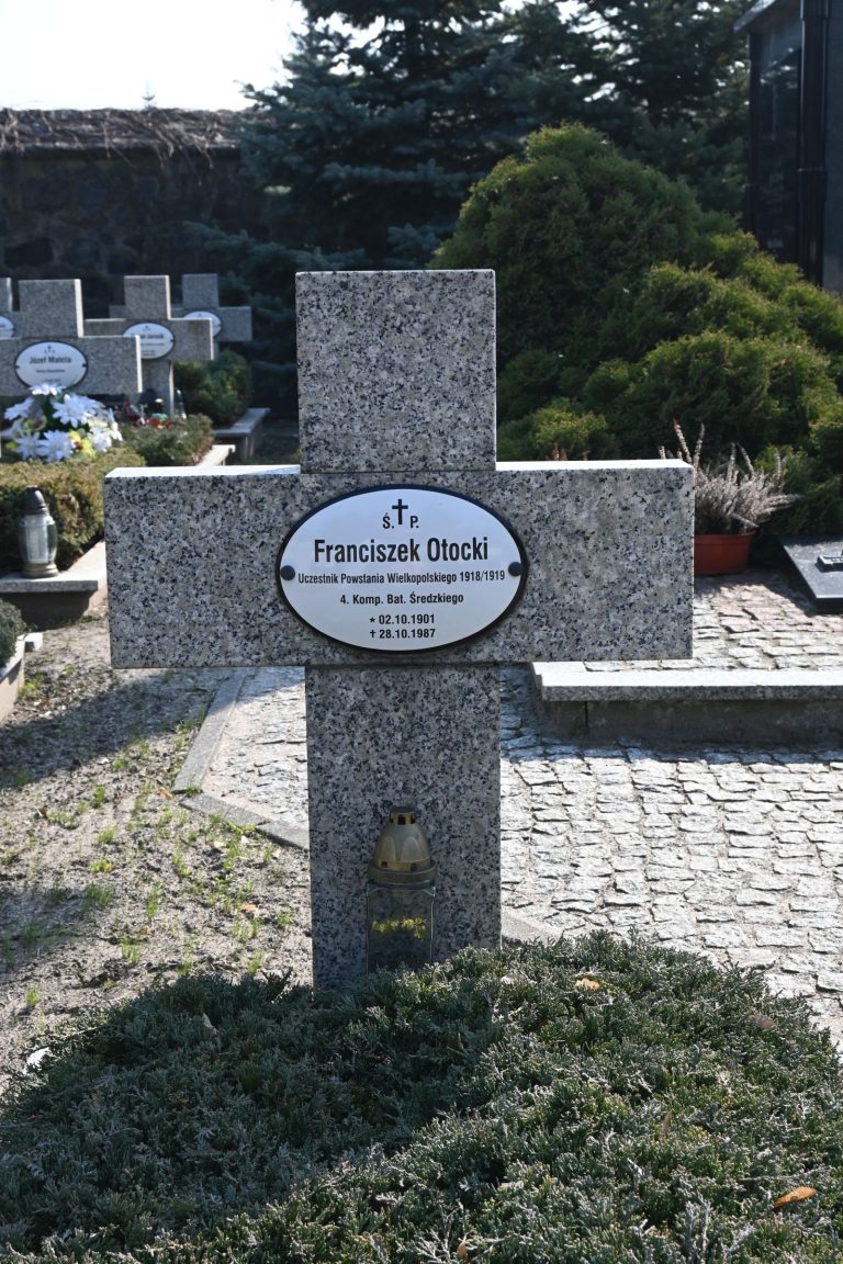 Kamienny krzyż nagrobny z owalną tabliczką "Franciszek Otocki"; na trzonie mosiężny uchwyt na znicz, u stóp gęsty jałowiec, w tle brukowana alejka i inne nagrobki.
