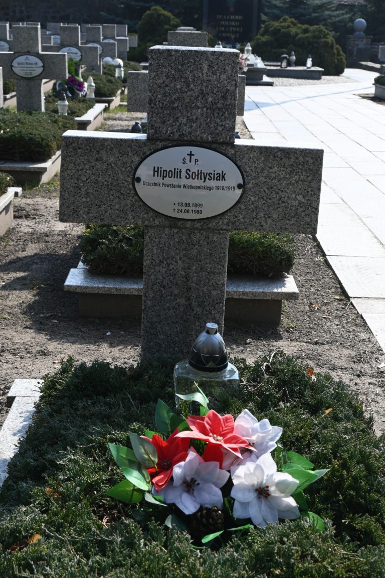 Kamienny krzyż nagrobny z owalną tabliczką "Hipolit Sołtysiak"; u stóp biało-czerwony bukiet z zielonymi liśćmi i szklany lampion, w tle alejka i rzędy nagrobków.