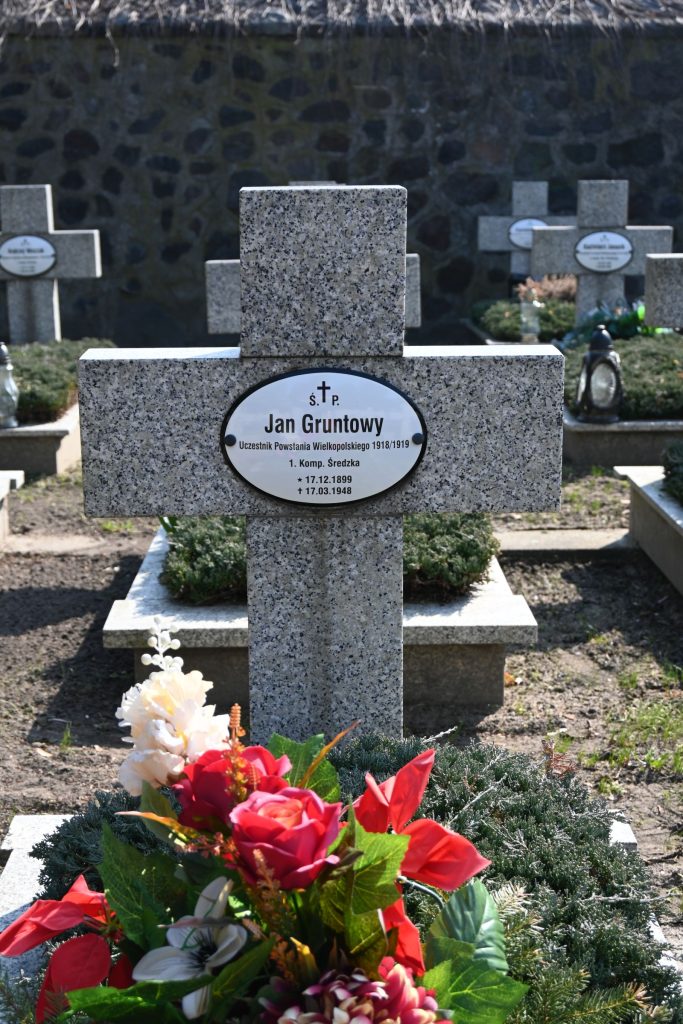 Kamienny krzyż nagrobny z owalną tabliczką „Jan Gruntowy”; przed krzyżem bukiet czerwonych i białych kwiatów na zielonym jałowcu, w tle kamienny mur i inne krzyże.