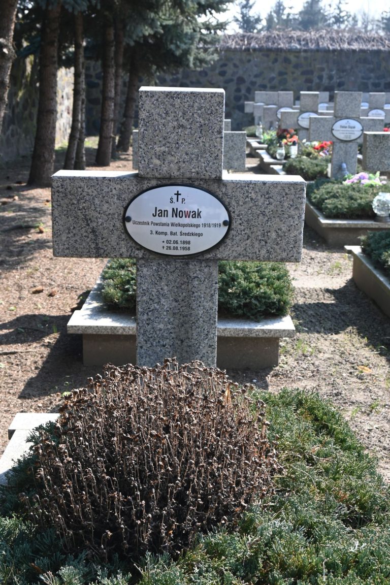 Kamienny krzyż nagrobny z owalną tabliczką „Jan Nowak”; przed krzyżem przycięty brązowy krzew na tle gęstego jałowca, w tle rzędy grobów i wysoki mur.