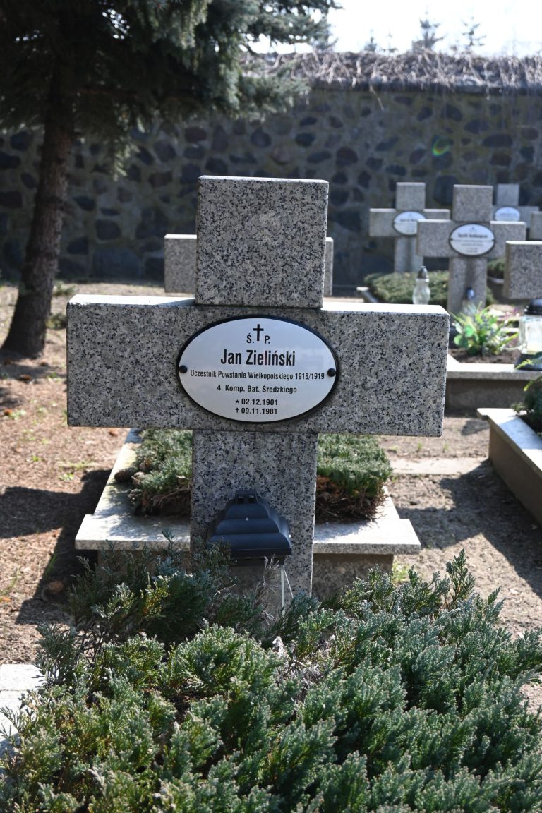 Kamienny krzyż nagrobny z owalną tabliczką „Jan Zieliński”; u podstawy gęsty jałowiec i czarny znicz, w tle kamienny mur i drzewo.