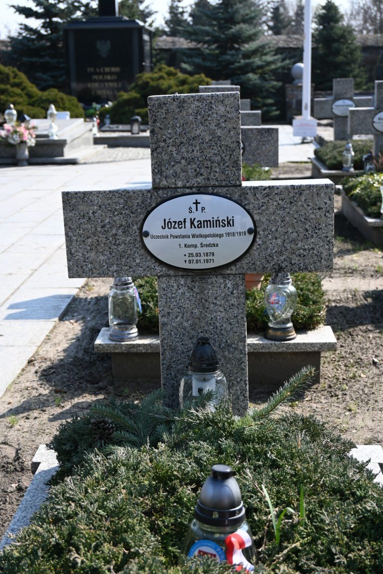 Kamienny krzyż z owalną tabliczką "Józef Kamiński"; u podstawy srebrny i czarny znicz oraz gałązki; po bokach dwa szklane lampiony z krzyżem; nagrobek otoczony niską zielenią, w tle alejka i inne groby.