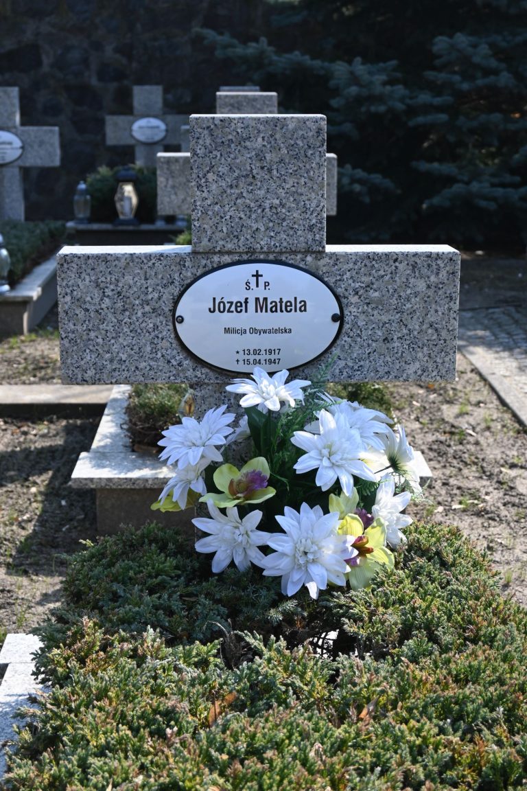 Kamienny krzyż nagrobny z owalną tabliczką „Józef Matela”; przed krzyżem bukiet dużych białych kwiatów z żółtymi środkami na jałowcu, w tle kamienny mur i drzewa.