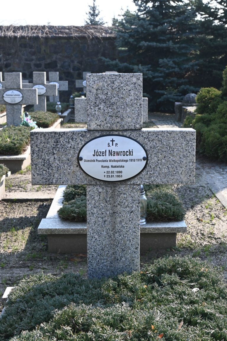 Kamienny krzyż nagrobny z owalną tabliczką „Józef Nawrocki”; u podstawy gęsty jałowiec, w tle rzędy grobów i kamienny mur.