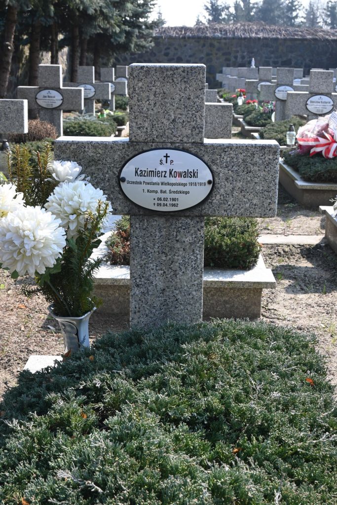 Kamienny krzyż nagrobny z owalną tabliczką „Kazimierz Kowalski”; obok krzyża duży biały bukiet chryzantem, u podstawy jałowiec, w tle rzędy nagrobków i murek.