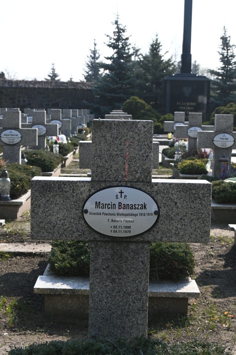 Kamienny krzyż nagrobny z owalną tabliczką „Marcin Banaszak”; na niskiej podstawie, otoczony zadbanymi krzewami jałowca, w tle murek i rzędy nagrobków.