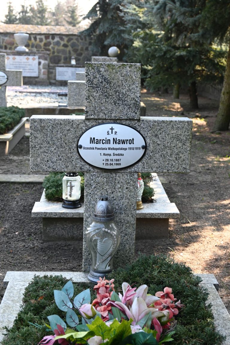 Kamienny krzyż z tabliczką "Marcin Nawrot"; u stóp duży srebrny znicz i szklana latarenka; w przodzie bukiet różowych lilii i zielonych liści; otoczony niską roślinnością, w tle inne krzyże i kamienny mur.