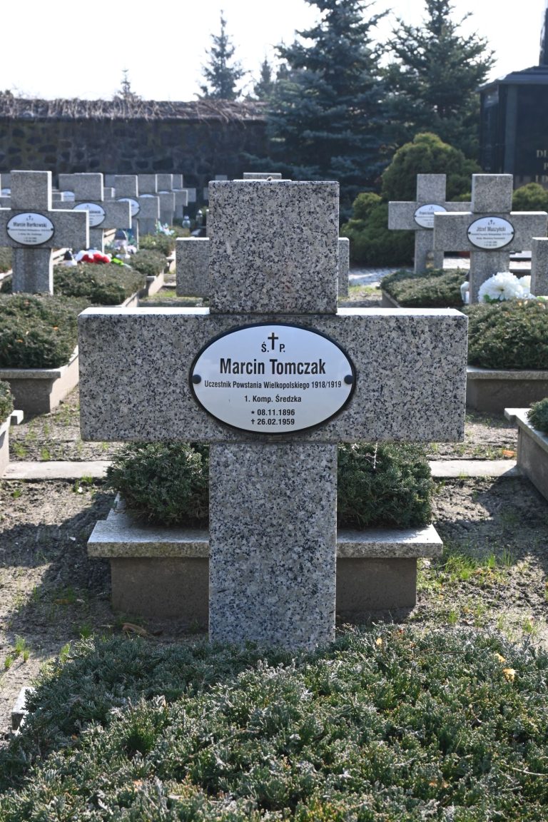 Kamienny krzyż nagrobny z owalną tabliczką „Marcin Tomczak”; u podstawy gęsty jałowiec, w tle rzędy nagrobków i drzew.