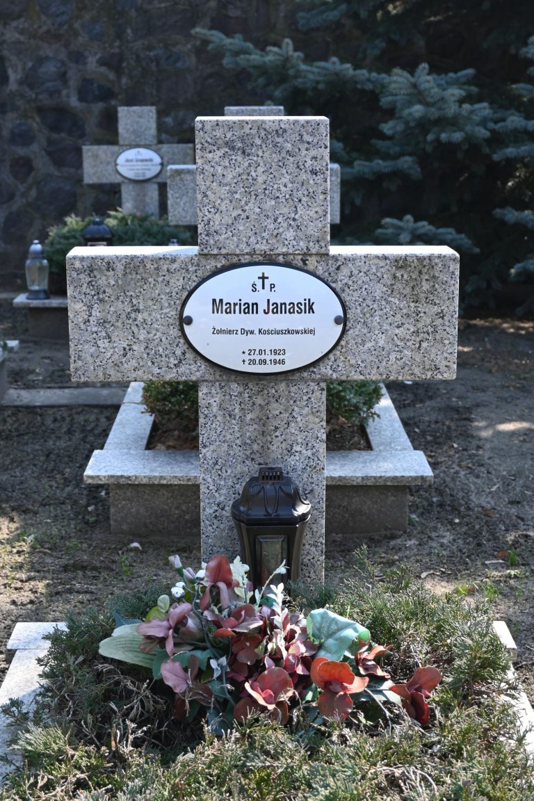 Kamienny krzyż z owalną tabliczką "Marian Janasik"; u podstawy czarny lampion i kompozycja zielonych liści z czerwonymi kwiatami; w tle kamienny mur i świerk.