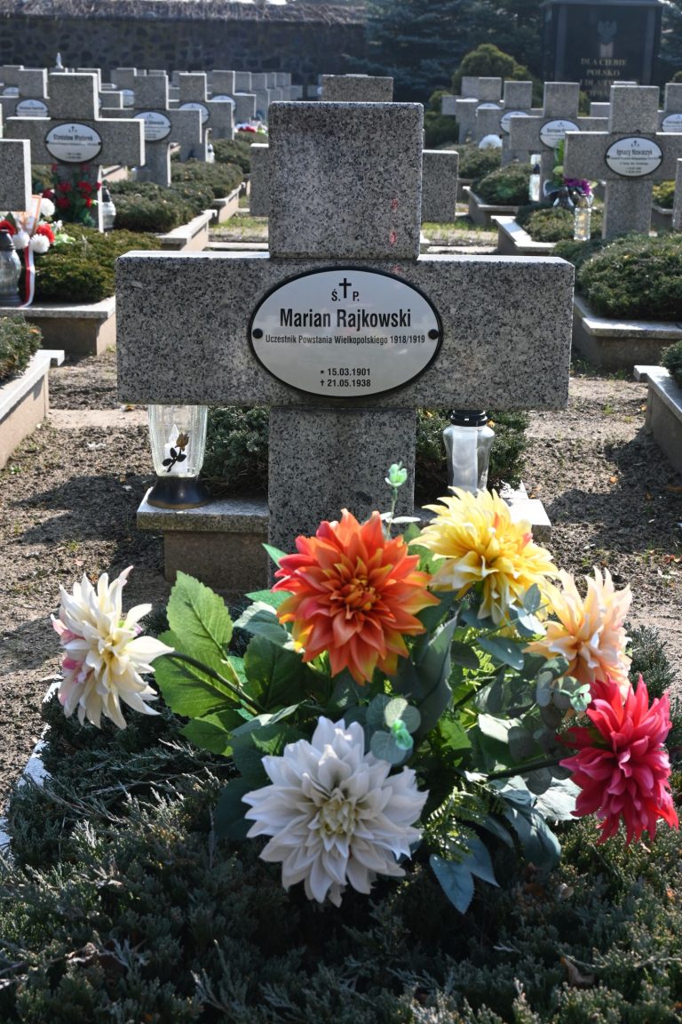 Kamienny krzyż z owalną tabliczką "Marian Rajkowski"; przed krzyżem wielobarwny bukiet dalii, po bokach szklane lampiony; w tle inne groby.