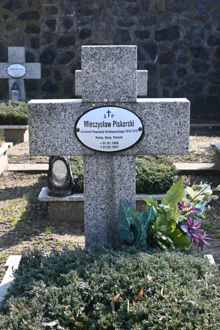 Kamienny krzyż z owalną tabliczką "Mieczysław Piskorski"; obok bukiet zielonych liści z fioletową lilią i szklana latarnia; tło stanowi kamienny mur.