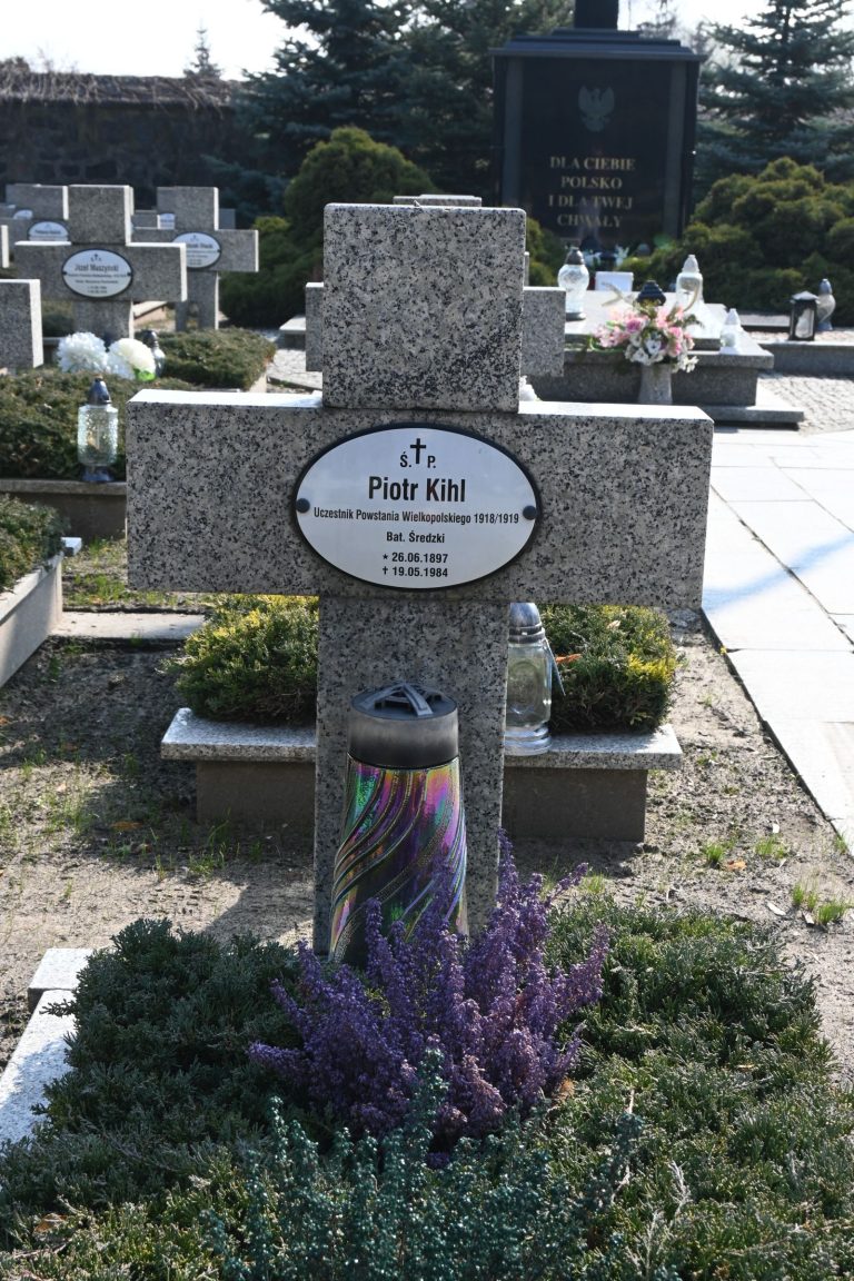 Kamienny krzyż z owalną tabliczką "Piotr Kihl"; u podstawy kolorowa świeca cylindryczna i kępa fioletowej wrzosowatej rośliny; w tle inne groby.