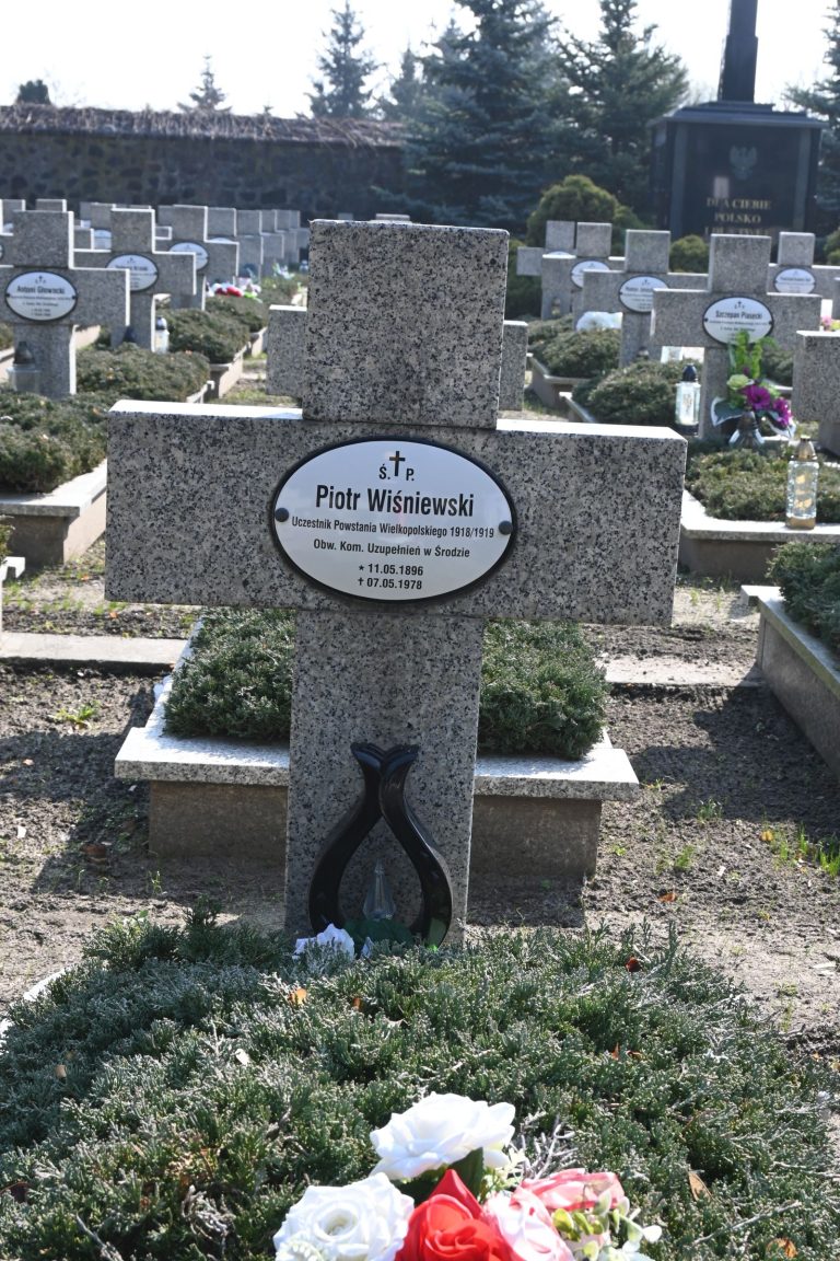 Kamienny krzyż z owalną tabliczką "Piotr Wiśniewski"; u podstawy czarny stojak w kształcie podkowy i bukiet biało-czerwonych kwiatów; w tle inne groby i czarny krzyż.