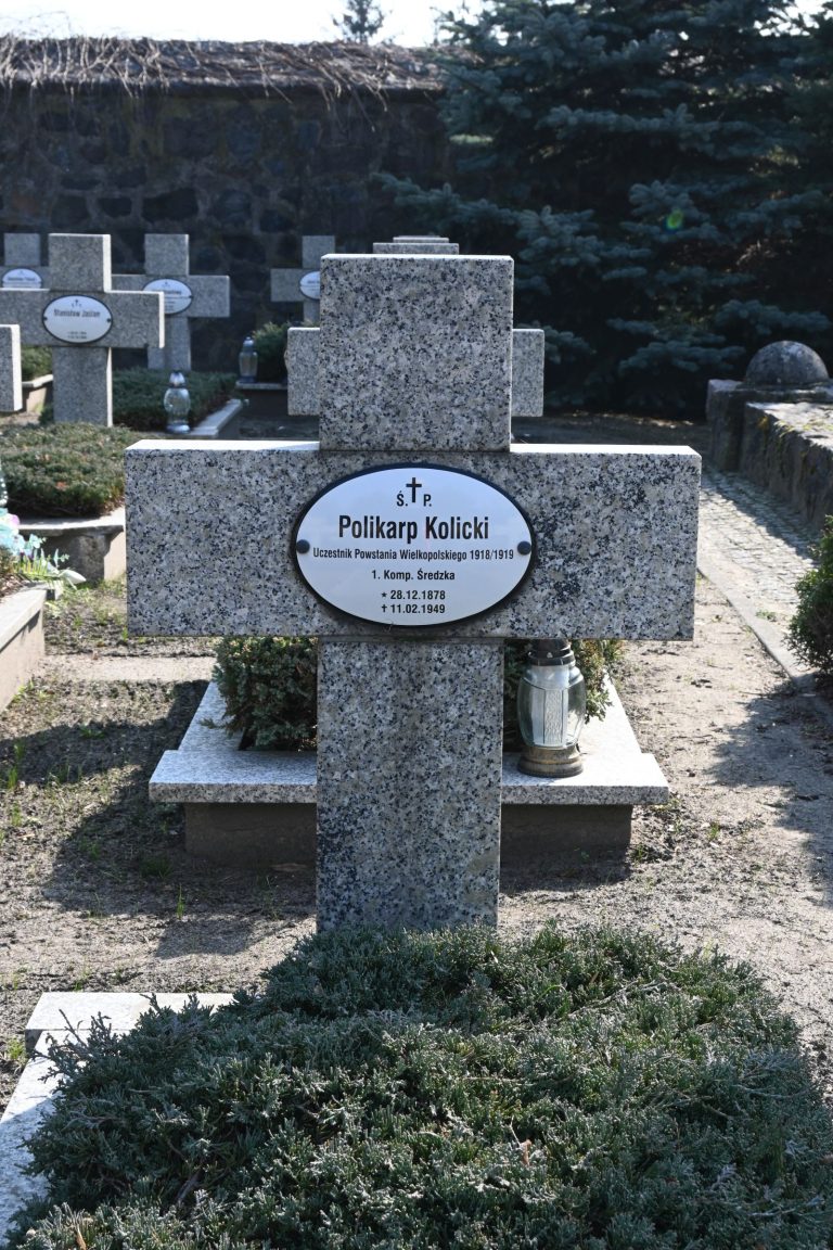 Kamienny krzyż z owalną tabliczką "Polikarp Kolicki"; obok szklana latarnia, u podstawy niski jałowiec; w tle mur z kamieni.
