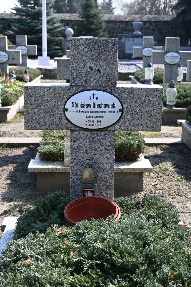 Kamienny krzyż z tabliczką "Stanisław Biechowiak" ustawiony na płycie grobu; u podstawy mała złota latarenka i gliniana donica, mogiła porośnięta niskimi krzewami; w tle rząd jednakowych krzyży i lampek.