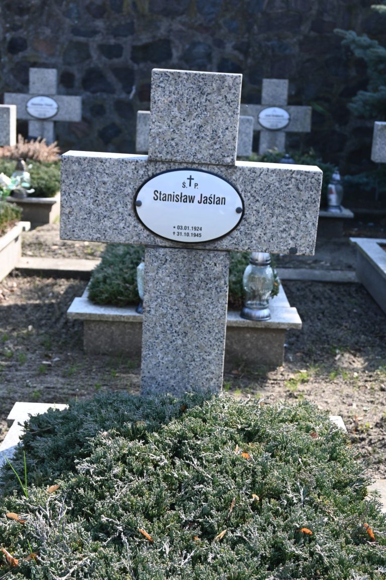 Nagrobek Stanisława Jaślana w Kwaterze Powstańców Wielkopolskich – betonowy krzyż z owalną tabliczką z imieniem; długa mogiła porośnięta jałowcem i metalowym zniczem; w tle rząd innych nagrobków.