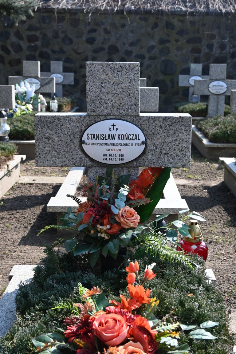 Nagrobek Stanisława Kończala w Kwaterze Powstańców Wielkopolskich – betonowy krzyż z owalną tabliczką z imieniem i nazwiskiem; przed nim barwny bukiet czerwonych i pomarańczowych kwiatów oraz zielona roślinność.