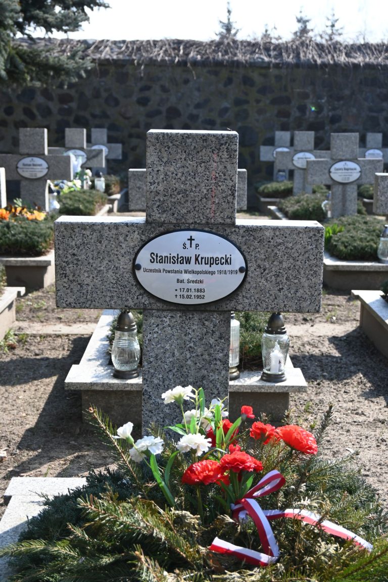 Nagrobek Stanisława Krupeckiego w Kwaterze Powstańców Wielkopolskich – kamienny krzyż z owalną tabliczką z imieniem i datami, flankowany dwiema metalowymi lampionami, u podstawy jałowiec z wieńcem z białych i czerwonych kwiatów udekorowanym wstążką.