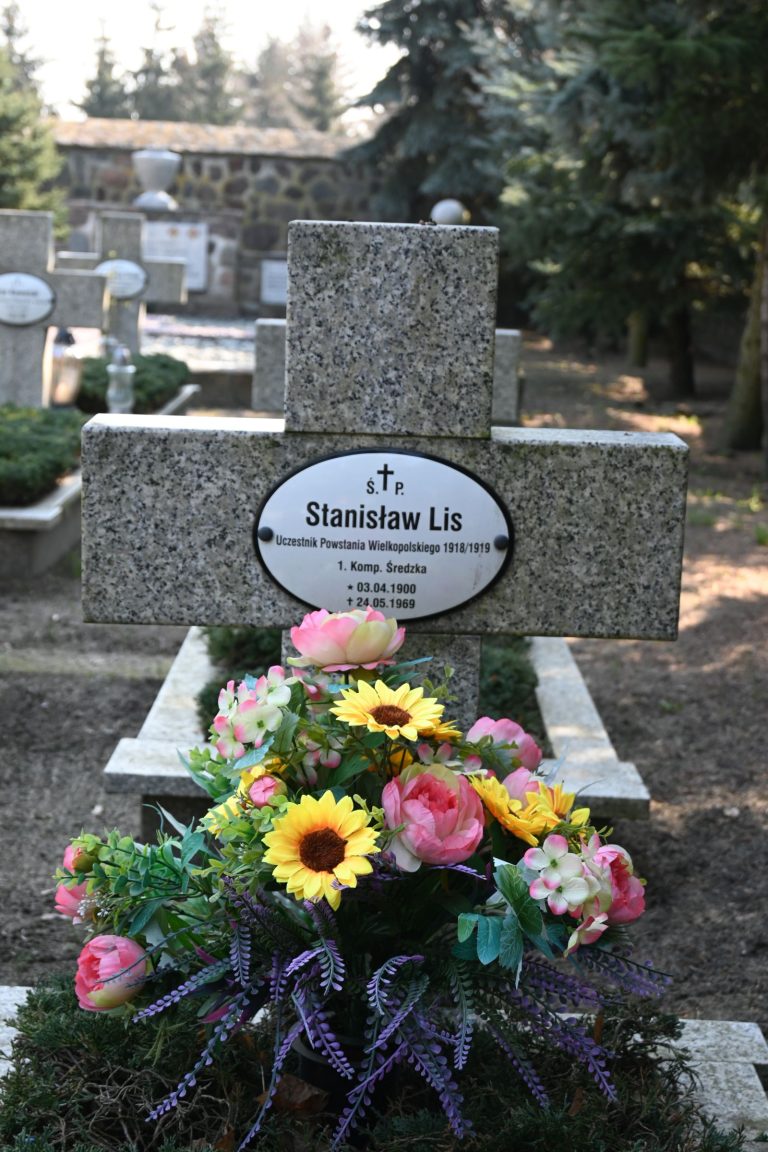 Krzyż z granitu z owalną tabliczką "Stanisław Lis", u podstawy barwna kompozycja kwiatów – słoneczniki, różowe piwonie i wrzosy; w tle mur z kamienia i inne groby.