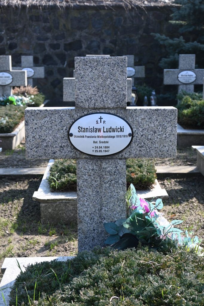 Nagrobek Stanisława Ludwickiego w Kwaterze Powstańców Wielkopolskich – betonowy krzyż z owalną tabliczką z imieniem, przed nim zielona wiązanka z różowymi kwiatami na długiej mogile porośniętej jałowcem.