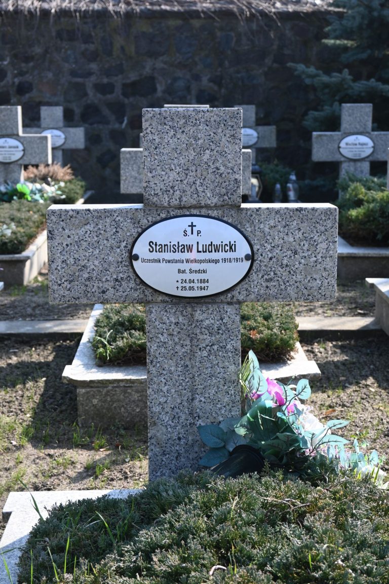 Nagrobek Stanisława Ludwickiego w Kwaterze Powstańców Wielkopolskich – betonowy krzyż z owalną tabliczką z imieniem, przed nim zielona wiązanka z różowymi kwiatami na długiej mogile porośniętej jałowcem.