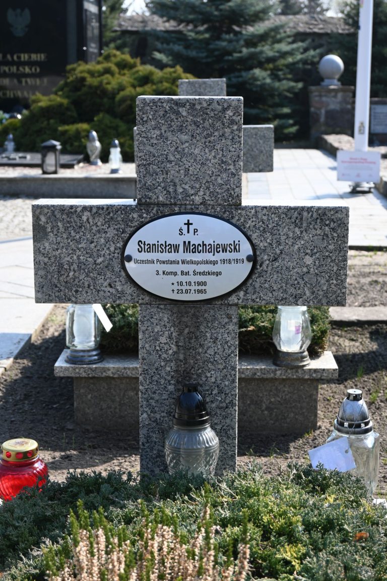 Nagrobek w formie kamiennego krzyża z owalną tabliczką "Stanisław Machajewski", otoczony wrzosem i zimozielonymi krzewami; u podstawy szare i czerwone znicze, w tle alejka i inne nagrobki.