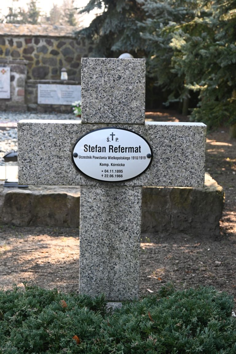 Kamienny krzyż z owalną tabliczką "Stefan Refermat", uczestnik Powstania Wielkopolskiego. U stóp krzyża rośnie niski, zielony krzew; w tle kamienny mur, drzewa i fragmenty innych nagrobków.