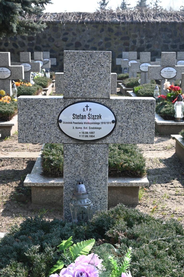 Nagrobek Stefana Ślazaka w Kwaterze Powstańców Wielkopolskich – betonowy krzyż z owalną tabliczką z imieniem, u podstawy metalowy znicz i bukiecik fioletowych kwiatów; w tle widoczne rzędy innych krzyży.