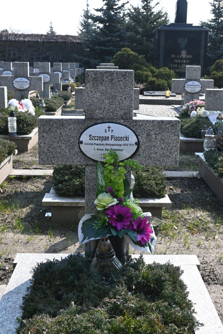 Nagrobek Szczepana Piaseckiego w Kwaterze Powstańców Wielkopolskich – betonowy krzyż z owalną tabliczką na nagrobku; przed krzyżem kolorowy bukiet zielonych i fioletowych kwiatów oraz metalowy znicz; w tle widoczne są inne mogiły i drzewo.