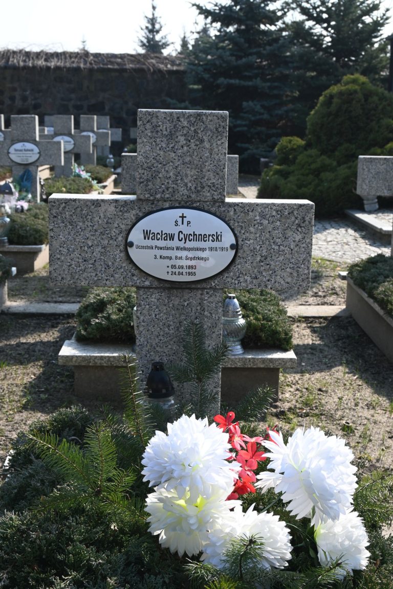 Nagrobek Wacława Cycherskiego w Kwaterze Powstańców Wielkopolskich – kamienny krzyż z owalną tabliczką z imieniem, biało-czerwone kwiaty i zielona roślinność u podstawy