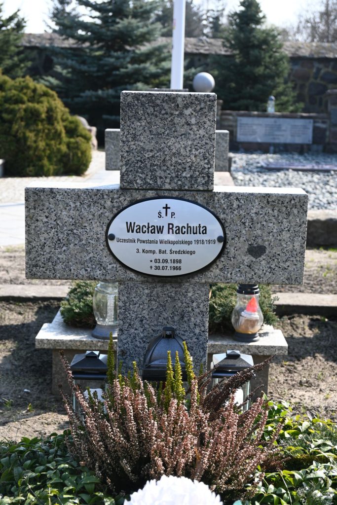Kamienny krzyż nagrobny z owalną tabliczką "Wacław Rachuta", uczestnik Powstania Wielkopolskiego. U stopy krzyża rośnie krzak wrzosu i ustawione są dwie metalowe latarenki; po bokach stoją znicze; w tle widoczne są inne groby i rośliny.