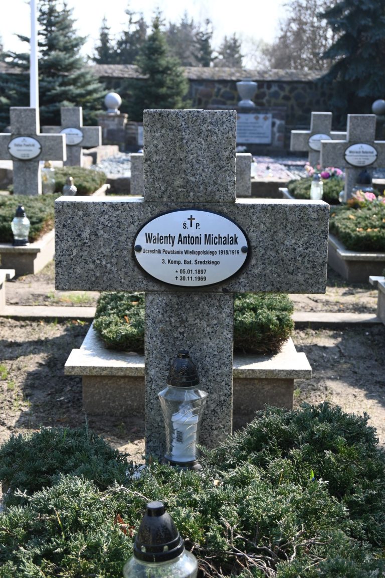 Kamienny krzyż z owalną tabliczką "Walenty Antoni Michalak" – uczestnik Powstania Wielkopolskiego; u podnóża krzyża biały szklany znicz i czarna latarenka, dookoła zielone krzewy; w tle rędy innych kamiennych krzyży i kamienny mur.