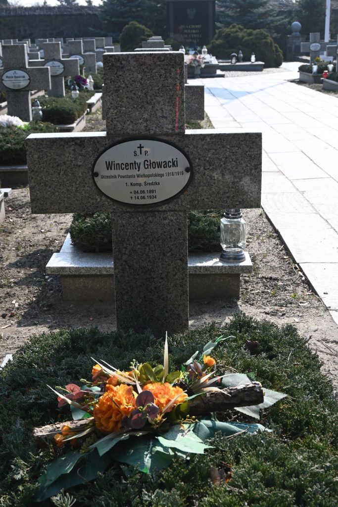 Nagrobek Wincentego Głowackiego w Kwaterze Powstańców Wielkopolskich – kamienny krzyż z owalną tabliczką z imieniem oraz bukiet pomarańczowo-zielonych kwiatów na mogile