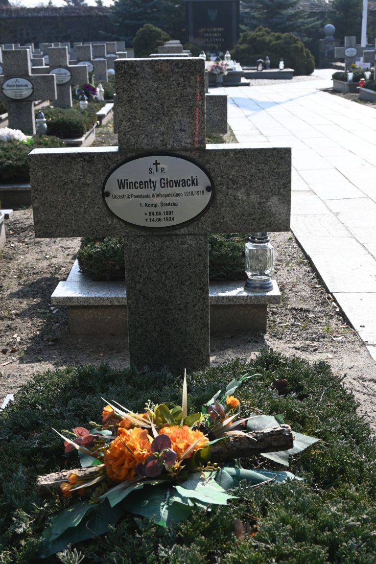 Nagrobek Wincentego Głowackiego w Kwaterze Powstańców Wielkopolskich – kamienny krzyż z owalną tabliczką z imieniem oraz bukiet pomarańczowo-zielonych kwiatów na mogile