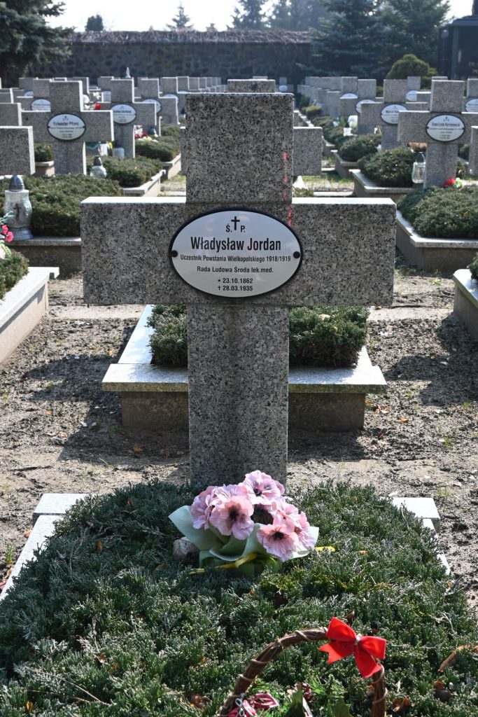 Nagrobek Władysława Jordana w Kwaterze Powstańców Wielkopolskich – kamienny krzyż z owalną tabliczką z imieniem i koszem różowych kwiatów u podstawy