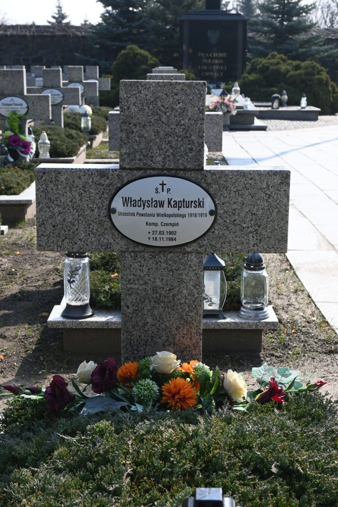 Nagrobek Władysława Kapturskiego w Kwaterze Powstańców Wielkopolskich – kamienny krzyż z owalną tabliczką z imieniem, kwiaty w kolorach żółtym, pomarańczowym i bordowym oraz znicze po obu stronach