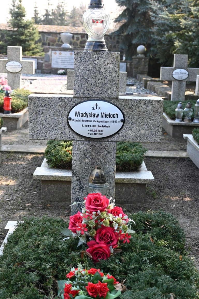 Nagrobek z kamiennym krzyżem i tabliczką "Władysław Mieloch", zwieńczony szklaną lampą i ozdobiony bukietem czerwonych i różowych kwiatów u podstawy.
