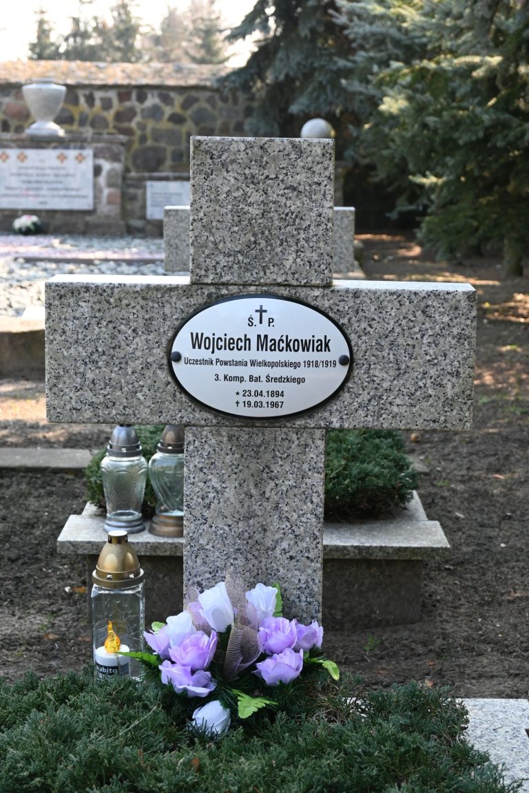 Nagrobek z krzyżem z tabliczką "Wojciech Maćkowiak", powstańca wielkopolskiego, ozdobiony białymi i fioletowymi kwiatami oraz kilkoma zniczami; w tle widoczny kamienny mur i inne groby.