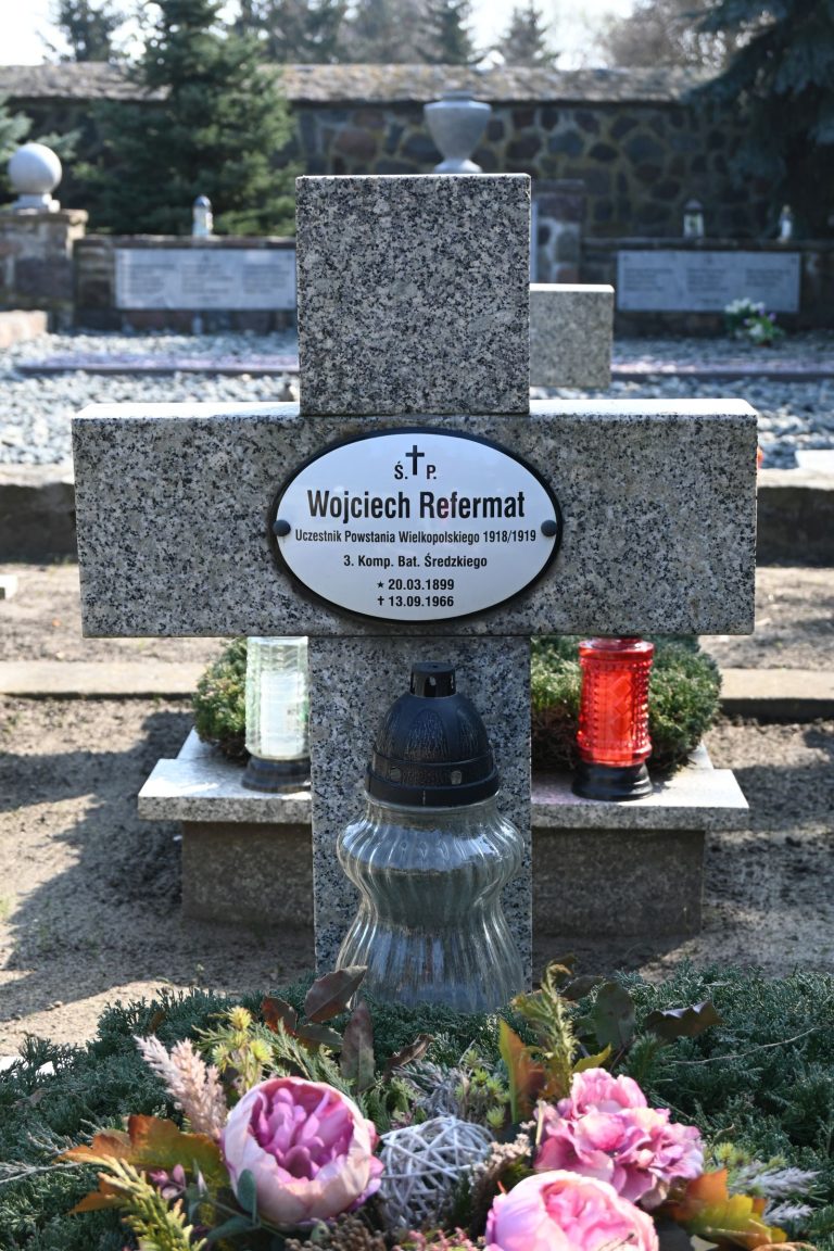 Zbliżenie na nagrobek z kamiennym krzyżem i tabliczką "Wojciech Refermat" oraz kolorowymi zniczami i różowymi kwiatami u podstawy.